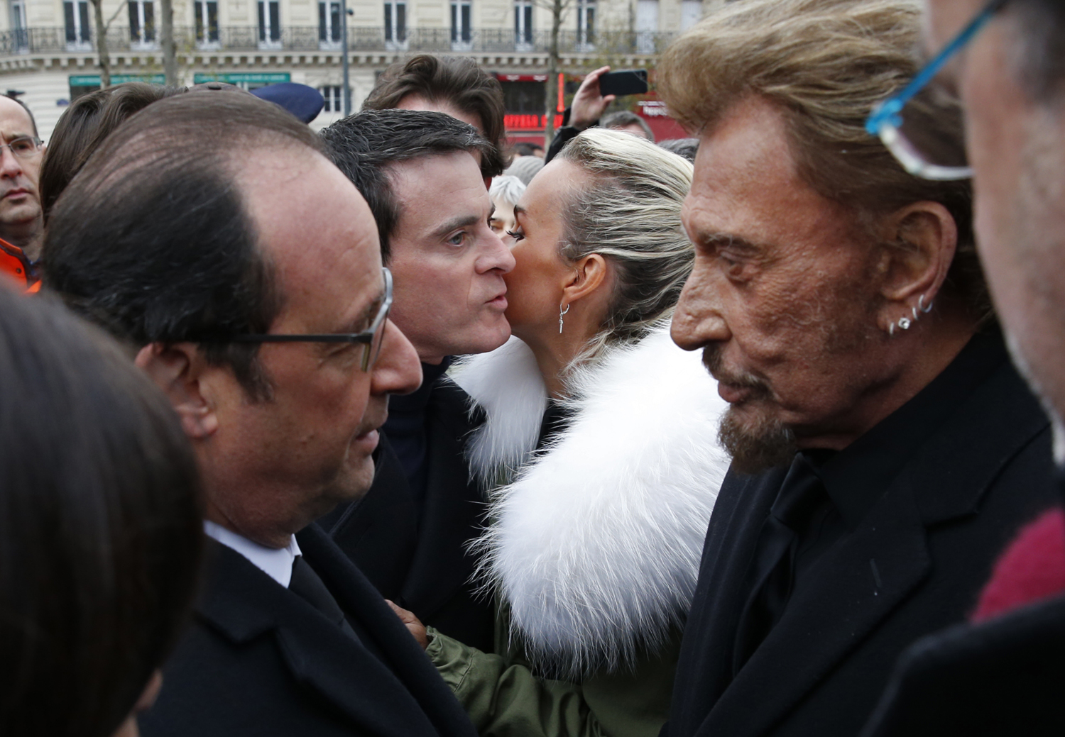 French President Francois Hollande (L) speaks with French singer Johnny Hallyday (R) as Prime Minister Manuel Valls (2ndL) kisses Hallyday's wife Laeticia at the end of a ceremony held on January 10, 2016 to mark a year since 1.6 million people thronged the French capital in a show of unity after attacks on the Charlie Hebdo newspaper and a Jewish supermarket. Just as it was last year, the vast Place de la Republique will be the focus of the gathering as people reiterate their support for freedom of expression and remember the other victims of what would become a year of jihadist outrages in France, culminating in the November 13 coordinated shootings and suicide bombings that killed 130 people and were claimed by the Islamic State (IS) group. / AFP / POOL / PHILIPPE WOJAZER