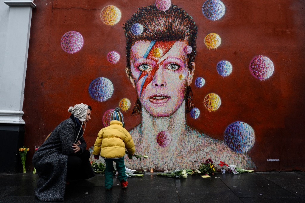 Floral tributes are left beneath a mural of British singer David Bowie, painted by Australian street artist James Cochran, aka Jimmy C, following the announcement of Bowie's death, in Brixton, south London, on January 11, 2016. British music icon David Bowie died of cancer at the age of 69, drawing an outpouring of tributes for the innovative star famed for groundbreaking hits like "Ziggy Stardust" and his theatrical shape-shifting style. AFP PHOTO / CHRIS RATCLIFFE RESTRICTED TO EDITORIAL USE, MANDATORY MENTION OF THE ARTIST UPON PUBLICATION, TO ILLUSTRATE THE EVENT AS SPECIFIED IN THE CAPTION / AFP / CHRIS RATCLIFFE
