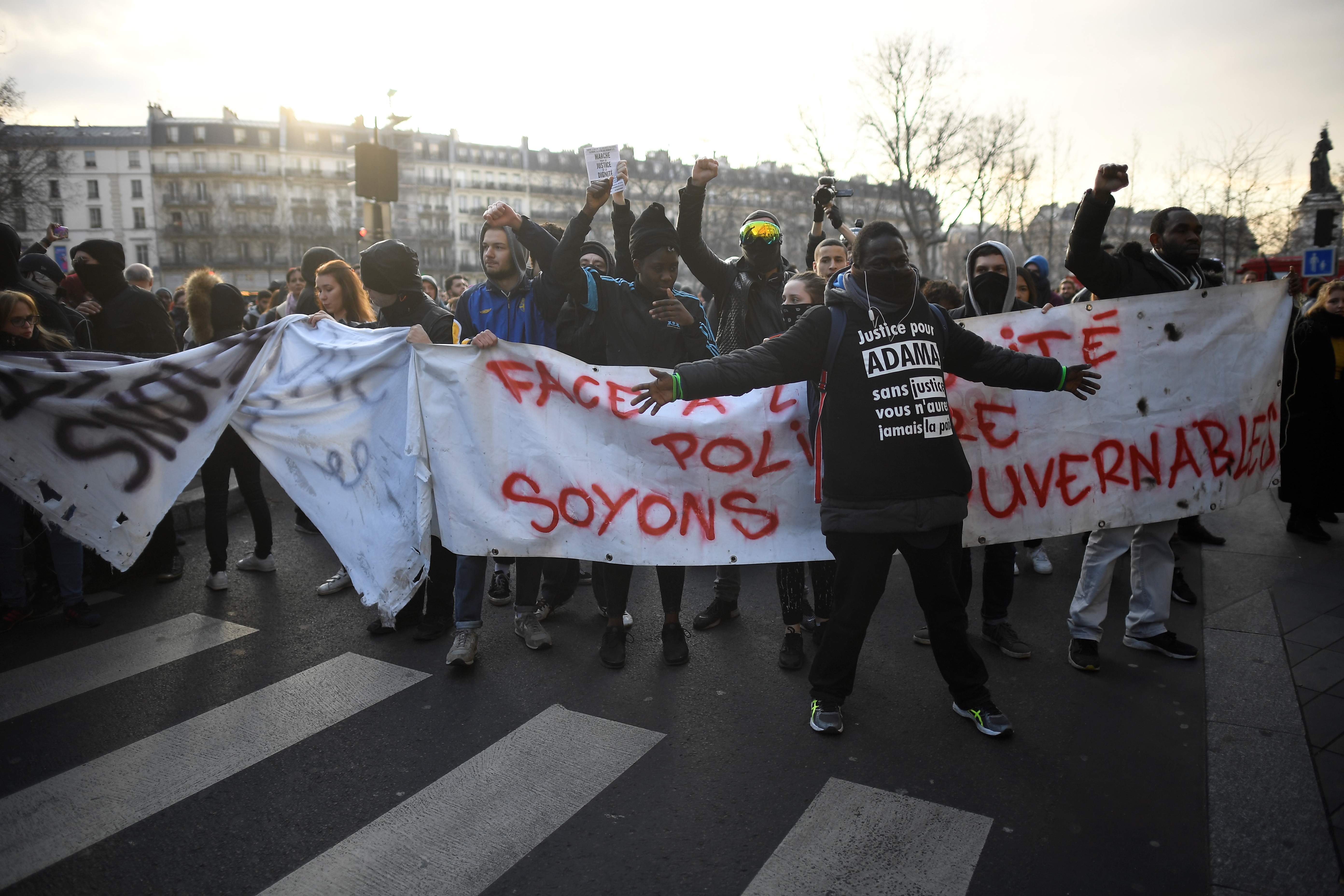 A protester wearing a shirt reading "Justice for Adama" gestures near a banner reading "In front of police impunity, let us be ungovernable" during a demonstration against police brutality on February 18, 2017 on the place de la Republique in Paris, following the alleged rape of a black youth, identified only as Theo, with a police baton, an incident that has sparked 10 nights of rioting and more than 200 arrests. The injuries sustained by Theo during a stop-and-search operation on February 2 in the suburb of Aulnay-sous-Bois, has revived long-simmering frustrations over policing in immigrant communities, where young men accuse the police of repeatedly targeting them in aggressive stop-and-search operations and using excessive force during arrests.  / AFP PHOTO / Lionel BONAVENTURE